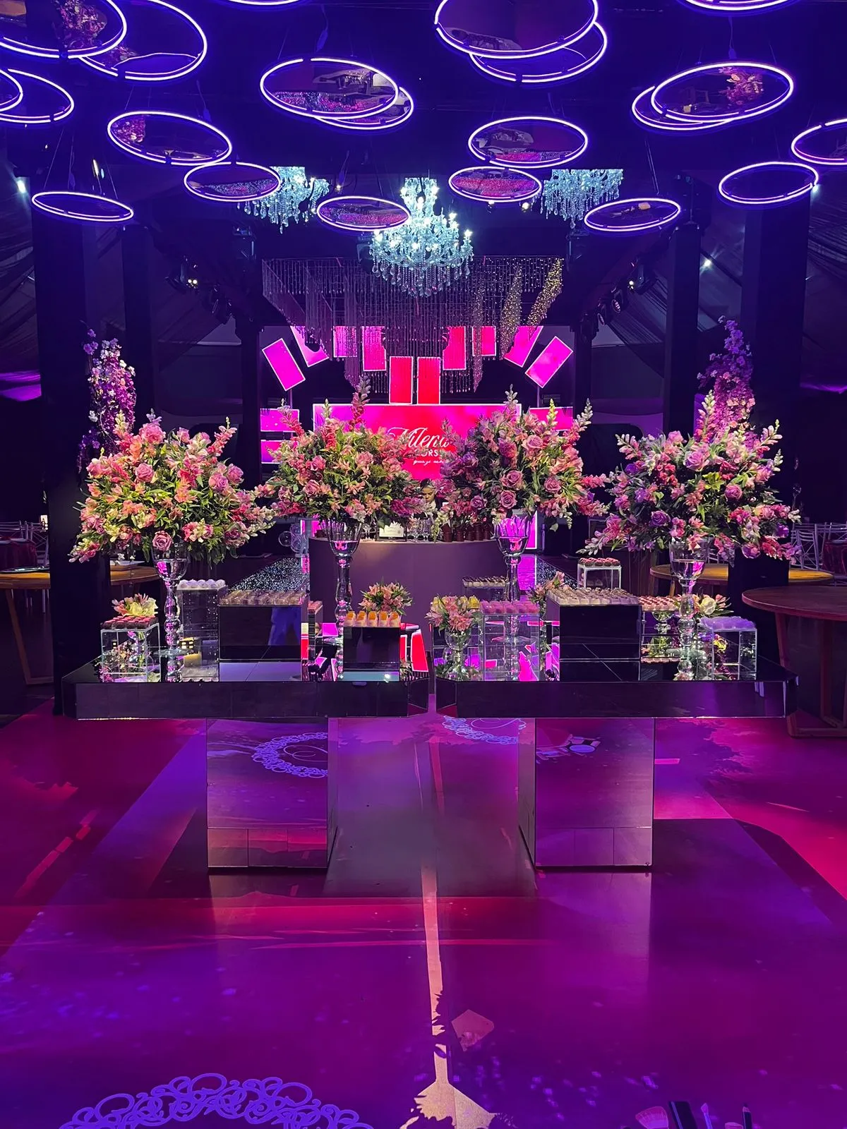Mesa de doces espelhada com arranjos florais e iluminação cênica em rosa