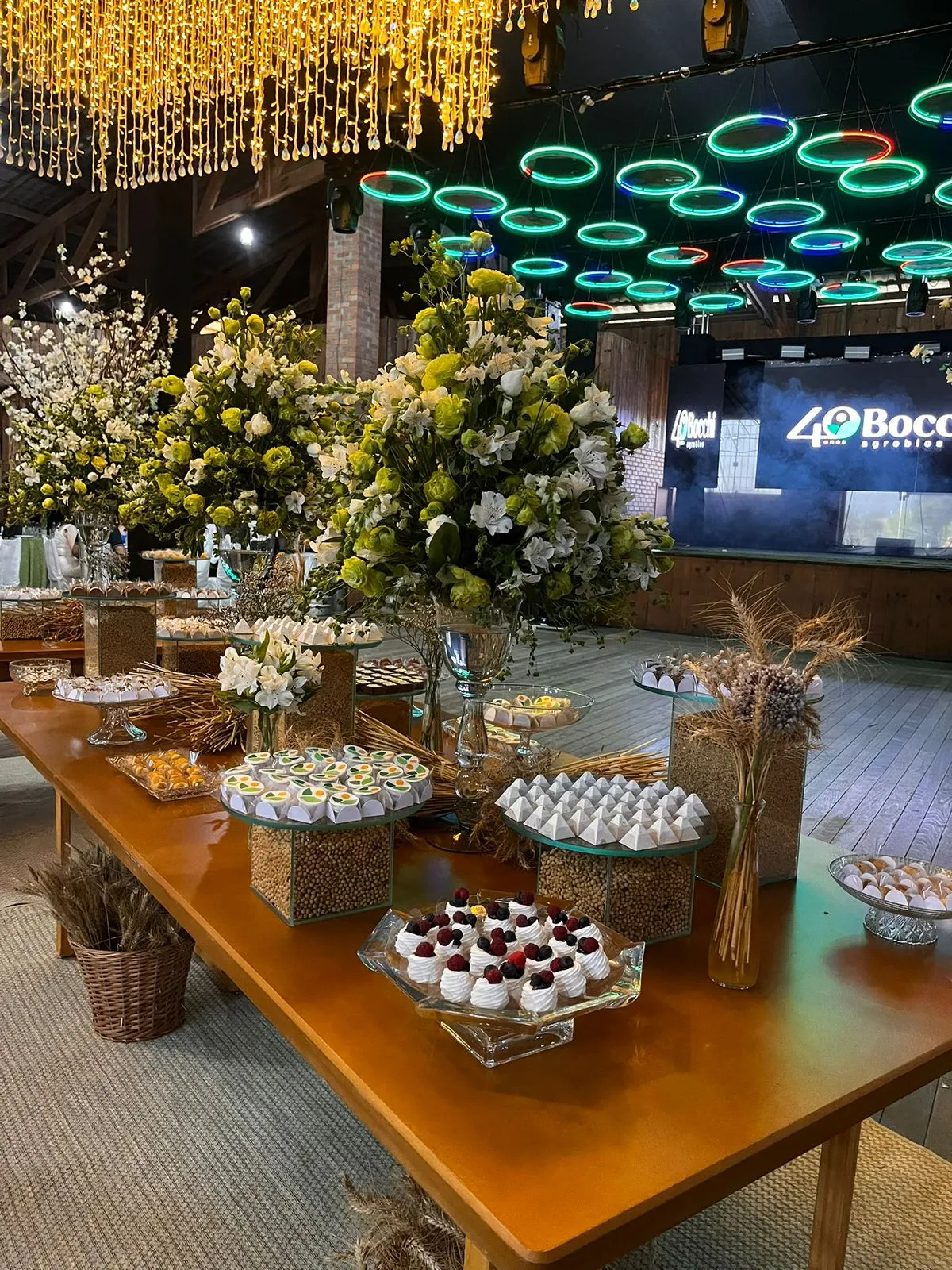 Mesa de doces de evento corporativo com arranjos florais e painel de LED ao fundo
