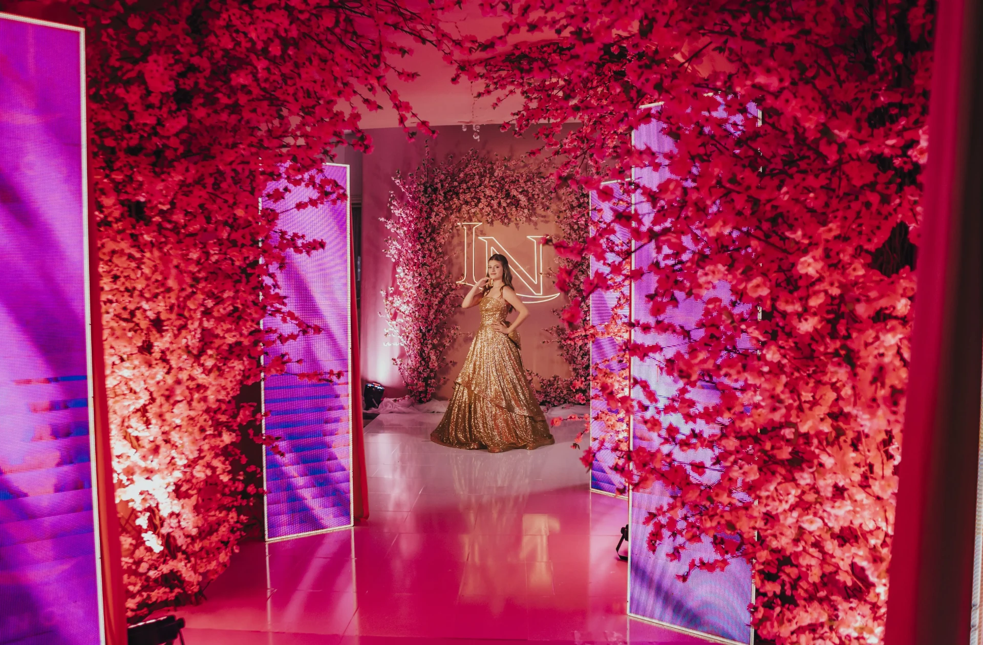 Debutante em vestido dourado na entrada decorada com cerejeiras e painéis de LED